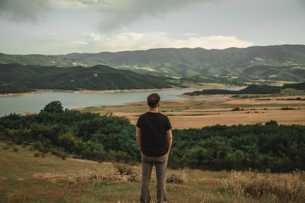 Homem contempla a paisagem de um lago e montanhas, simbolizando a conquista de uma vida plena e a atitude de gratidão pelas coisas simples.