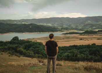 Homem contempla a paisagem de um lago e montanhas, simbolizando a conquista de uma vida plena e a atitude de gratidão pelas coisas simples.