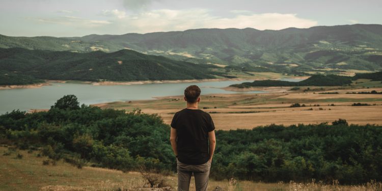 Homem contempla a paisagem de um lago e montanhas, simbolizando a conquista de uma vida plena e a atitude de gratidão pelas coisas simples.