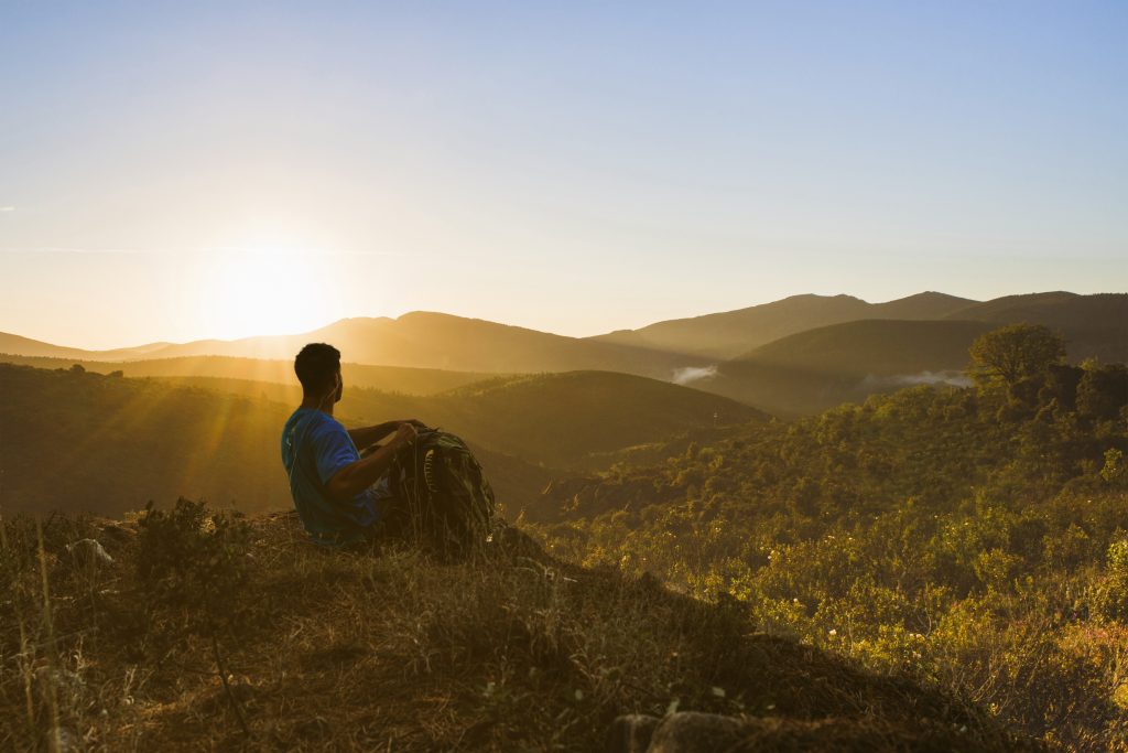 Jovem sentado no topo de uma colina em um momento de autoconhecimento, observando o pôr do sol, simbolizando o início de uma jornada para transformar a vida.