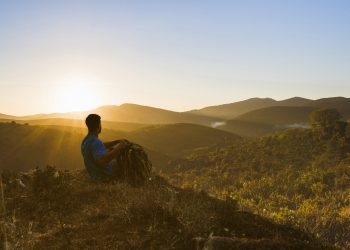 Jovem sentado no topo de uma colina em um momento de autoconhecimento, observando o pôr do sol, simbolizando o início de uma jornada para transformar a vida.