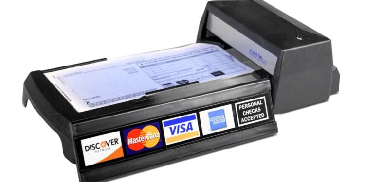 Máquina decalcadora de cartão de crédito manual, conhecida como "knuckle-buster", sobre um fundo branco. O aparelho é preto e mostra os logotipos da Discover, MasterCard, Visa e American Express.