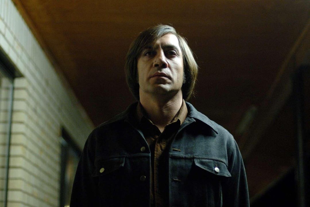 Anton Chigurh em cena icônica do filme "Onde os Fracos Não Têm Vez", encarando a câmera sob iluminação sombria em um corredor de motel.