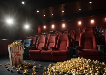 Sala de cinema vazia com poltronas vermelhas, vista do corredor. Em primeiro plano, um balde de pipoca clássico e pipoca derramada no chão, com a luz do projetor ao fundo.
