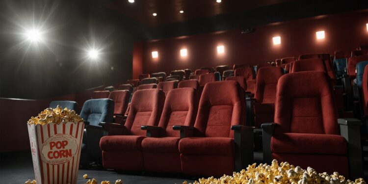 Sala de cinema vazia com poltronas vermelhas, vista do corredor. Em primeiro plano, um balde de pipoca clássico e pipoca derramada no chão, com a luz do projetor ao fundo.