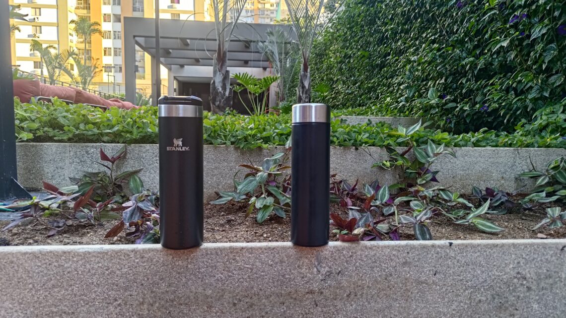 Comparativo visual entre a garrafa térmica Stanley Aerolight preta, à esquerda, e uma garrafa térmica de aço SUS304 também preta, à direita. As duas estão sobre um muro de concreto com um jardim ao fundo e são o objeto do teste sobre o gosto da água descrito no artigo.