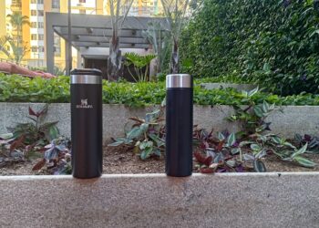 Comparativo visual entre a garrafa térmica Stanley Aerolight preta, à esquerda, e uma garrafa térmica de aço SUS304 também preta, à direita. As duas estão sobre um muro de concreto com um jardim ao fundo e são o objeto do teste sobre o gosto da água descrito no artigo.