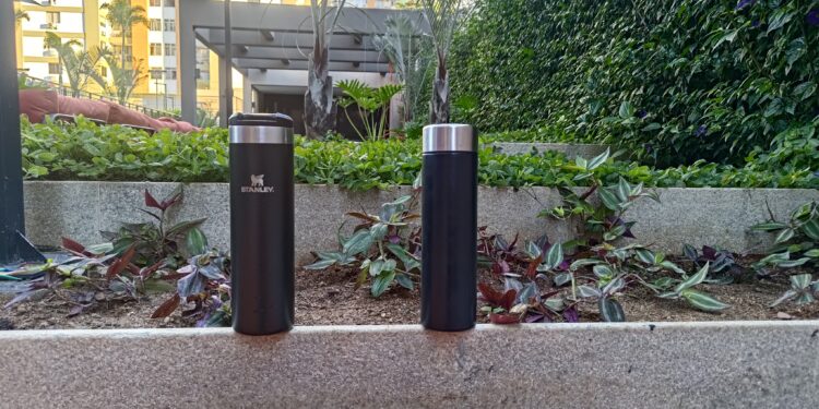 Comparativo visual entre a garrafa térmica Stanley Aerolight preta, à esquerda, e uma garrafa térmica de aço SUS304 também preta, à direita. As duas estão sobre um muro de concreto com um jardim ao fundo e são o objeto do teste sobre o gosto da água descrito no artigo.