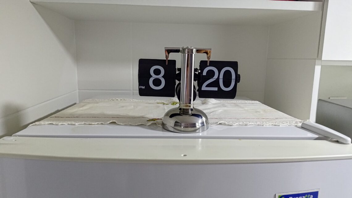 Relógio de mesa estilo flip clock retrô, com números pretos e estrutura prateada, posicionado em cima de um móvel branco.