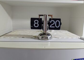 Relógio de mesa estilo flip clock retrô, com números pretos e estrutura prateada, posicionado em cima de um móvel branco.