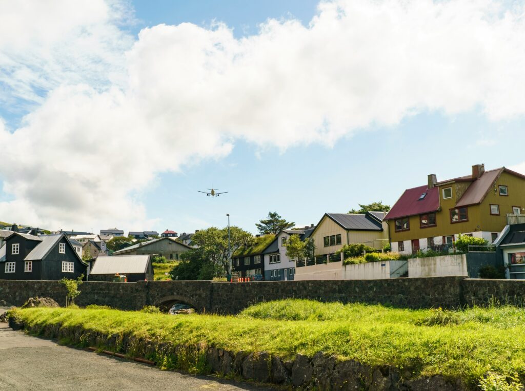 Uma aeronave sobrevoa um bairro residencial com casas de telhados coloridos e gramados verdes, evocando a sensação de voltar para casa e revisitar lugares familiares da infância.