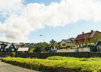 Uma aeronave sobrevoa um bairro residencial com casas de telhados coloridos e gramados verdes, evocando a sensação de voltar para casa e revisitar lugares familiares da infância.