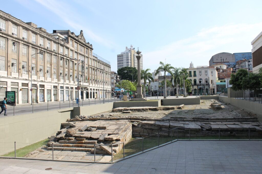 Vista do sítio arqueológico do Cais do Valongo, no Rio de Janeiro, com as ruínas de pedra em primeiro plano e os prédios da Zona Portuária ao fundo.