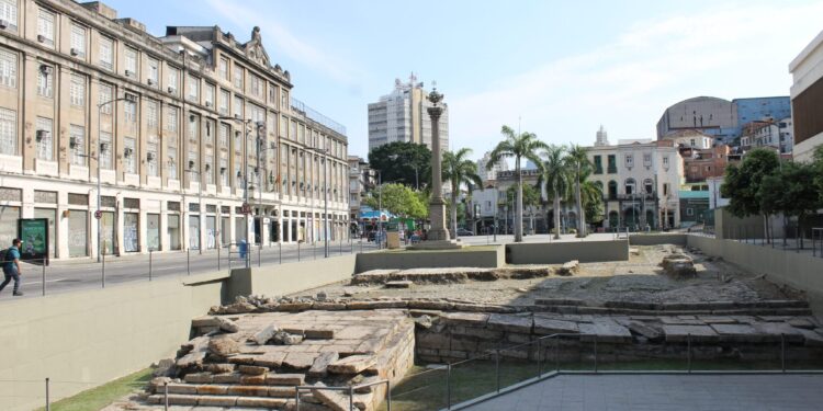 Vista do sítio arqueológico do Cais do Valongo, no Rio de Janeiro, com as ruínas de pedra em primeiro plano e os prédios da Zona Portuária ao fundo.