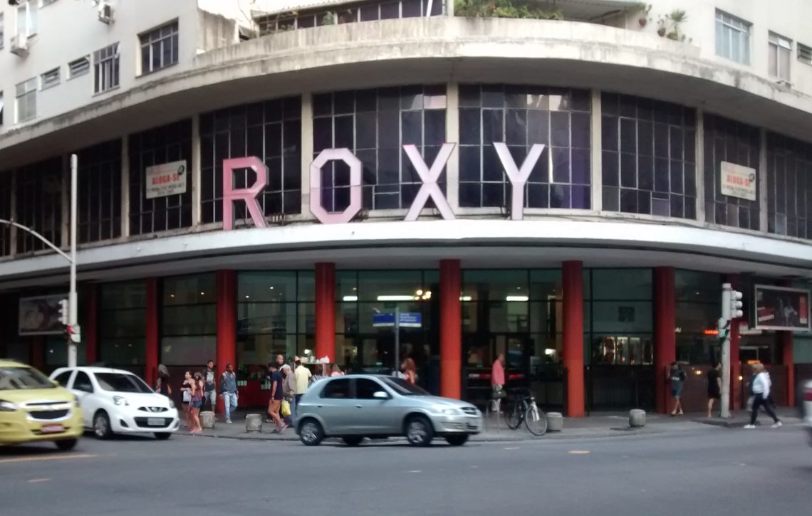 Fachada do antigo Cinema Roxy em Copacabana, Rio de Janeiro, com seu letreiro icônico cor-de-rosa e a movimentação de pedestres e carros na rua.