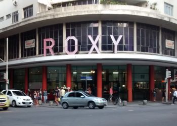 Fachada do antigo Cinema Roxy em Copacabana, Rio de Janeiro, com seu letreiro icônico cor-de-rosa e a movimentação de pedestres e carros na rua.