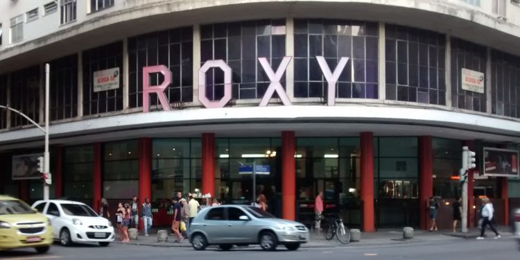Fachada do antigo Cinema Roxy em Copacabana, Rio de Janeiro, com seu letreiro icônico cor-de-rosa e a movimentação de pedestres e carros na rua.