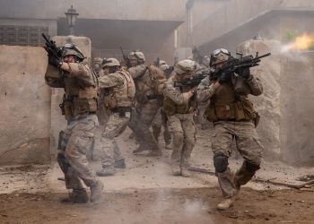 Soldados em uniforme de combate avançam por uma rua em ruínas durante um tiroteio, em uma cena de ação do filme "Warfare".