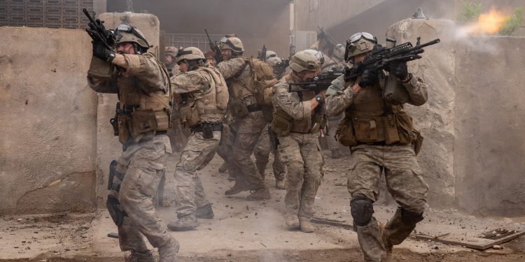 Soldados em uniforme de combate avançam por uma rua em ruínas durante um tiroteio, em uma cena de ação do filme "Warfare".