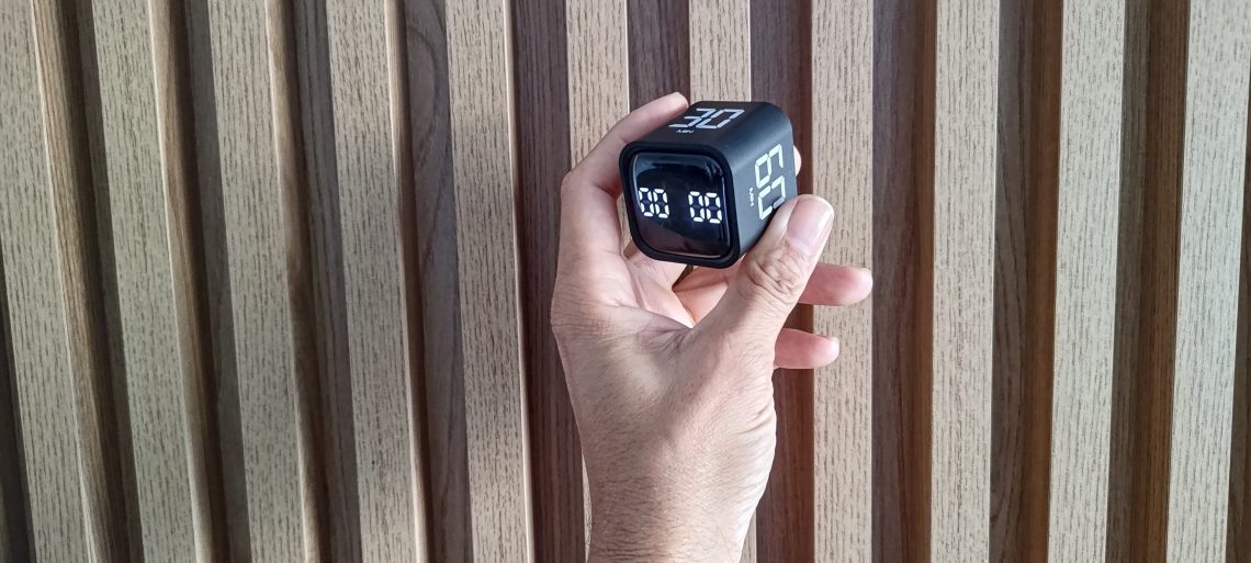 Mão de um homem segurando um timer cubo preto contra um fundo de painel de madeira canelada, ilustrando o gadget de produtividade.