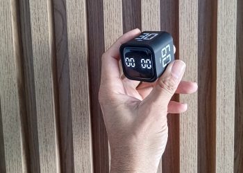 Mão de um homem segurando um timer cubo preto contra um fundo de painel de madeira canelada, ilustrando o gadget de produtividade.