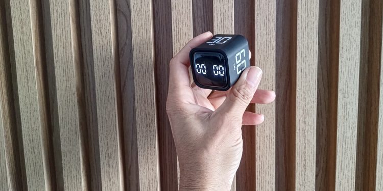 Mão de um homem segurando um timer cubo preto contra um fundo de painel de madeira canelada, ilustrando o gadget de produtividade.