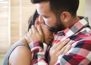 Um homem abraça uma mulher, confortando-a. Ele segura seu rosto com as mãos, demonstrando apoio e carinho em um momento de tristeza ou dificuldade.