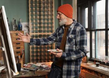 Homem mais velho, de barba e gorro laranja, sorrindo enquanto pinta um quadro em seu estúdio, representando a alegria de trabalhar com propósito.