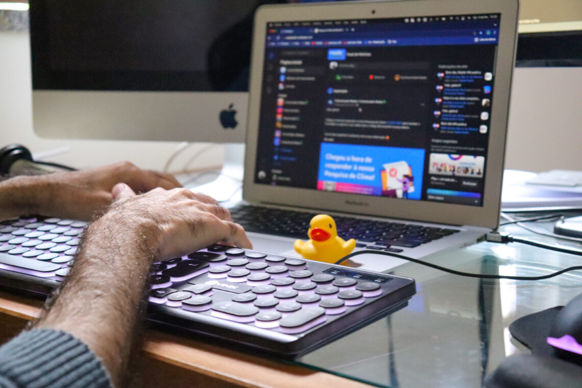 Homem digitando em um teclado com um pato de borracha amarelo em cima de um notebook, fazendo alusão ao aplicativo Kwack que emite sons de pato.