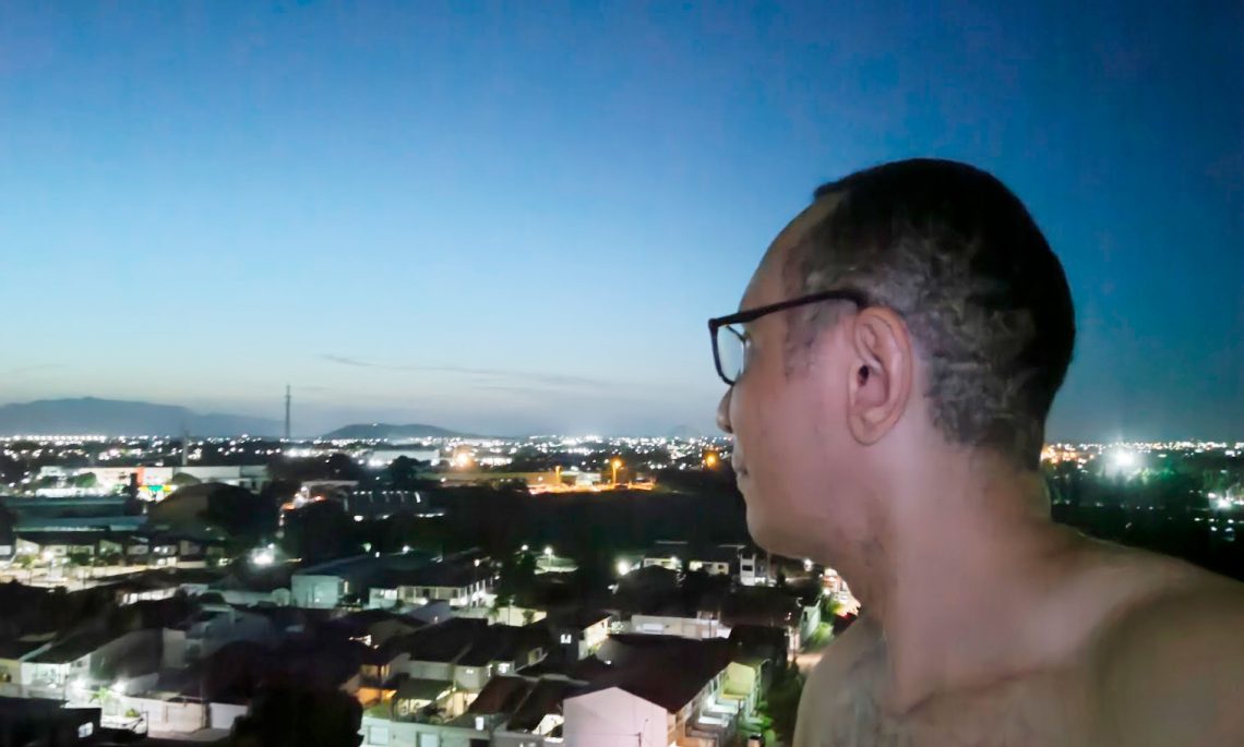 Homem de óculos, em perfil, olhando para o horizonte de uma cidade ao anoitecer, em um momento de reflexão sobre a jornada de autoconhecimento.