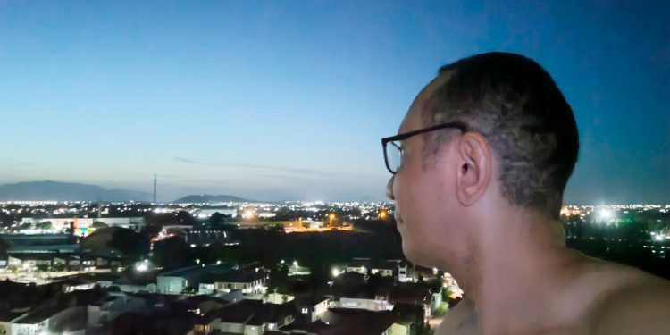 Homem de óculos, em perfil, olhando para o horizonte de uma cidade ao anoitecer, em um momento de reflexão sobre a jornada de autoconhecimento.