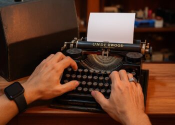 Close-up das mãos de um homem digitando em uma máquina de escrever Underwood preta e antiga sobre uma mesa de madeira.