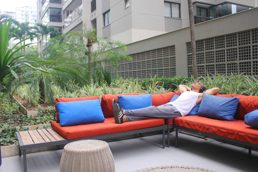 Homem deitado em um sofá laranja em uma área externa, relaxando com as mãos na cabeça enquanto ouve algo com fones de ouvido, ilustrando o consumo de audiolivros.