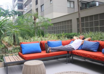 Homem deitado em um sofá laranja em uma área externa, relaxando com as mãos na cabeça enquanto ouve algo com fones de ouvido, ilustrando o consumo de audiolivros.