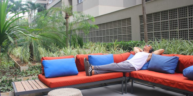 Homem deitado em um sofá laranja em uma área externa, relaxando com as mãos na cabeça enquanto ouve algo com fones de ouvido, ilustrando o consumo de audiolivros.