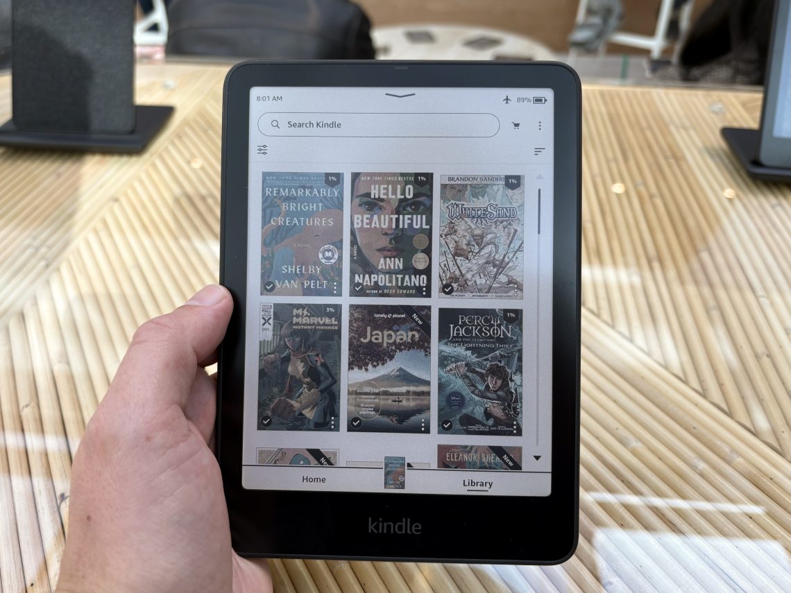 Mão segurando o novo Kindle Colorido, exibindo a biblioteca com capas de livros coloridas na tela.