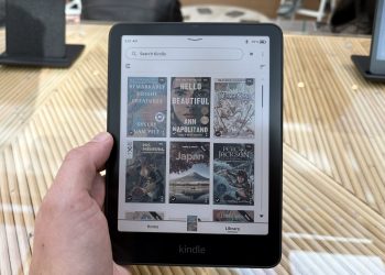 Mão segurando o novo Kindle Colorido, exibindo a biblioteca com capas de livros coloridas na tela.