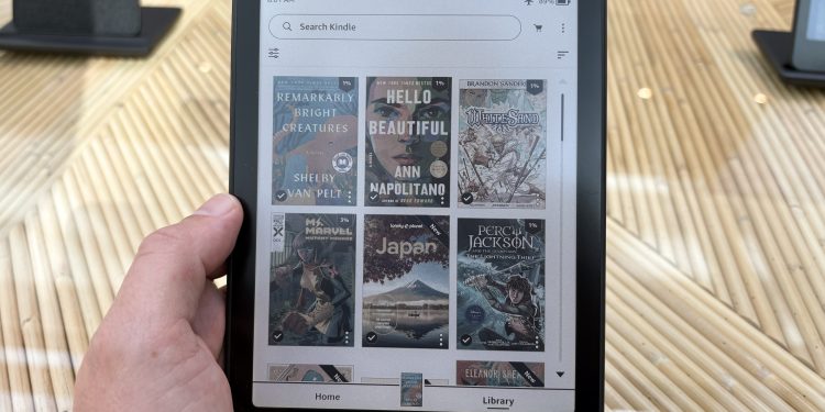 Mão segurando o novo Kindle Colorido, exibindo a biblioteca com capas de livros coloridas na tela.