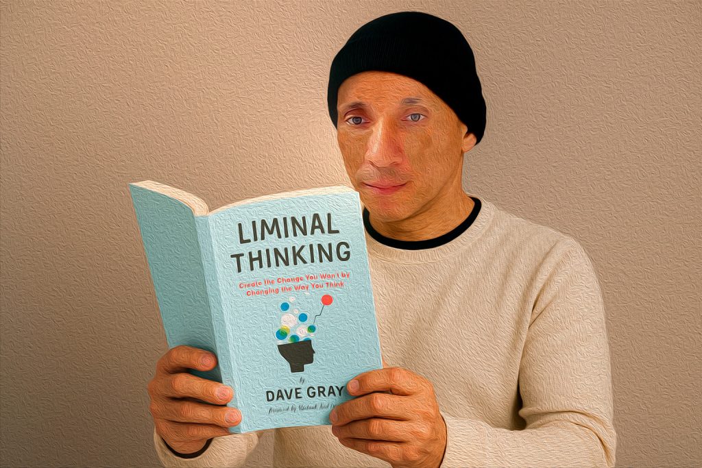 Homem de touca preta e blusa clara lê atentamente o livro 'Liminal Thinking' de Dave Gray, segurando-o com as duas mãos em frente a um fundo neutro.