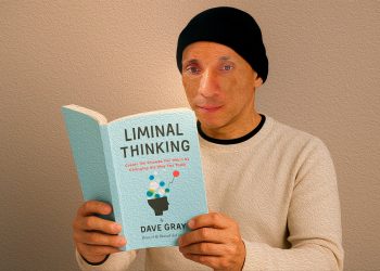 Homem de touca preta e blusa clara lê atentamente o livro 'Liminal Thinking' de Dave Gray, segurando-o com as duas mãos em frente a um fundo neutro.