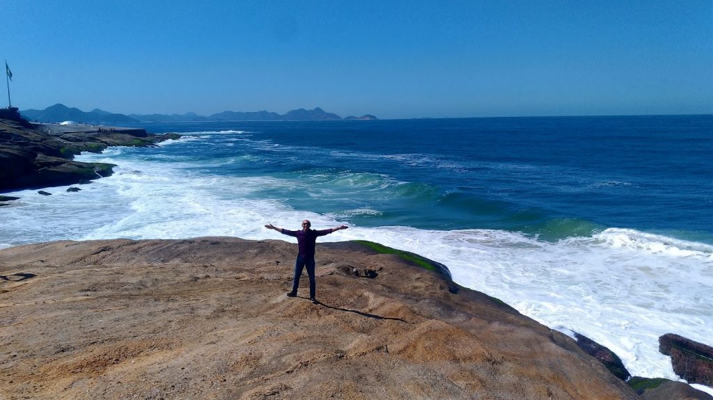 Gleydson, o homem de braços abertos em uma grande pedra na praia, com o mar azul e montanhas ao fundo, simbolizando a liberdade discutida no artigo.