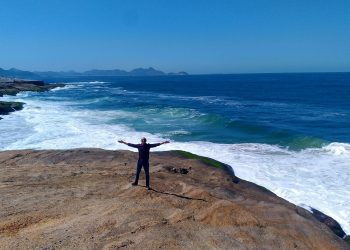 Gleydson, o homem de braços abertos em uma grande pedra na praia, com o mar azul e montanhas ao fundo, simbolizando a liberdade discutida no artigo.
