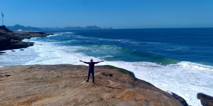 Gleydson, o homem de braços abertos em uma grande pedra na praia, com o mar azul e montanhas ao fundo, simbolizando a liberdade discutida no artigo.