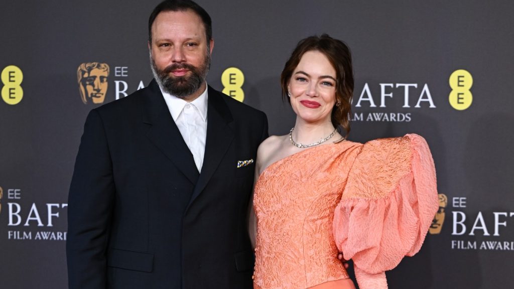 O diretor Yorgos Lanthimos, de terno preto e barba, posa ao lado da atriz Emma Stone, que veste um vestido laranja de ombro único, no tapete vermelho do BAFTA Awards.