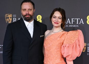 O diretor Yorgos Lanthimos, de terno preto e barba, posa ao lado da atriz Emma Stone, que veste um vestido laranja de ombro único, no tapete vermelho do BAFTA Awards.