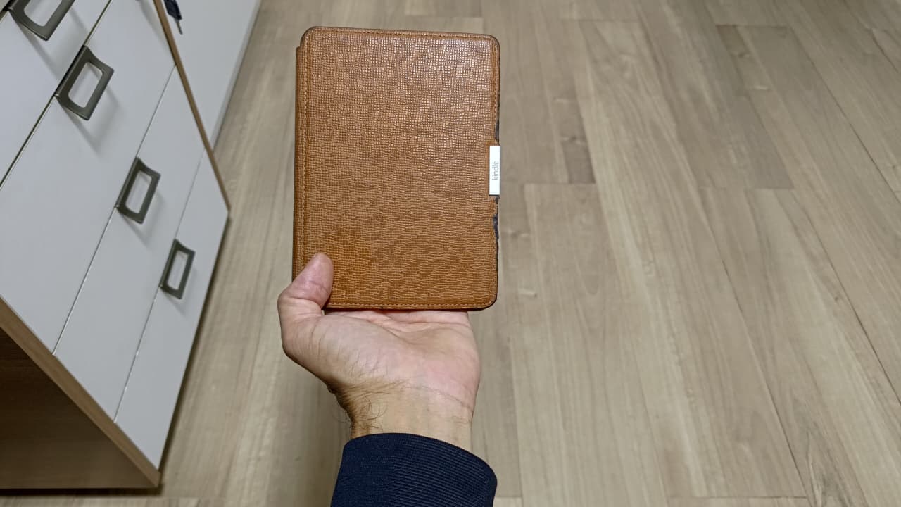 Mão segurando um Kindle antigo com capa de couro marrom, representando o modelo Paperwhite que ainda funciona bem.