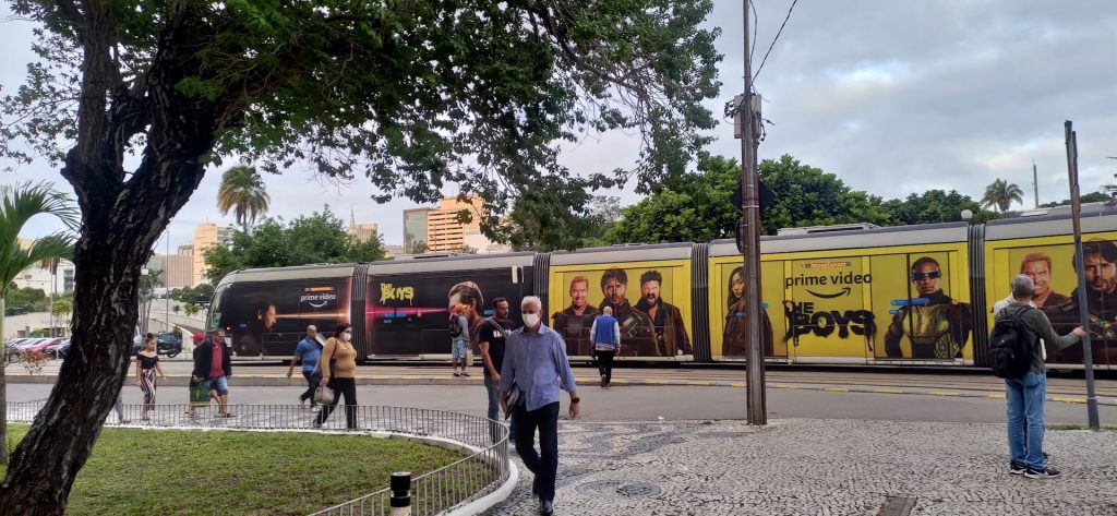 VLT no Rio de Janeiro com anúncio da série The Boys, enquanto pessoas caminham pela calçada em um fim de tarde, representando o palco de histórias do cotidiano.