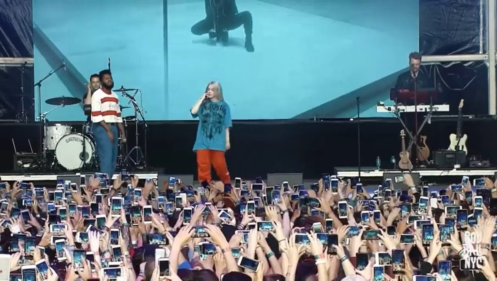 Billie Eilish no palco vista de frente, com uma multidão abaixo dela gravando o show com seus celulares erguidos, criando um mar de telas.