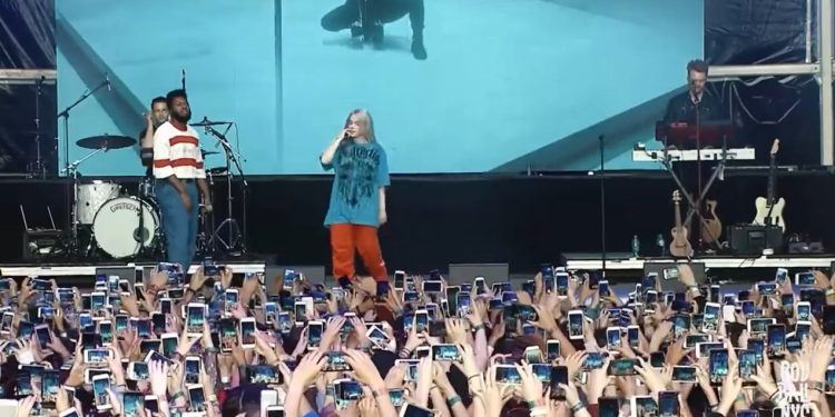 Billie Eilish no palco vista de frente, com uma multidão abaixo dela gravando o show com seus celulares erguidos, criando um mar de telas.