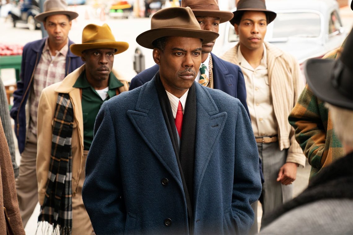 Chris Rock como o personagem Loy Cannon na quarta temporada da série Fargo, usando um casaco azul e chapéu, com outros homens ao fundo em uma cena de rua.